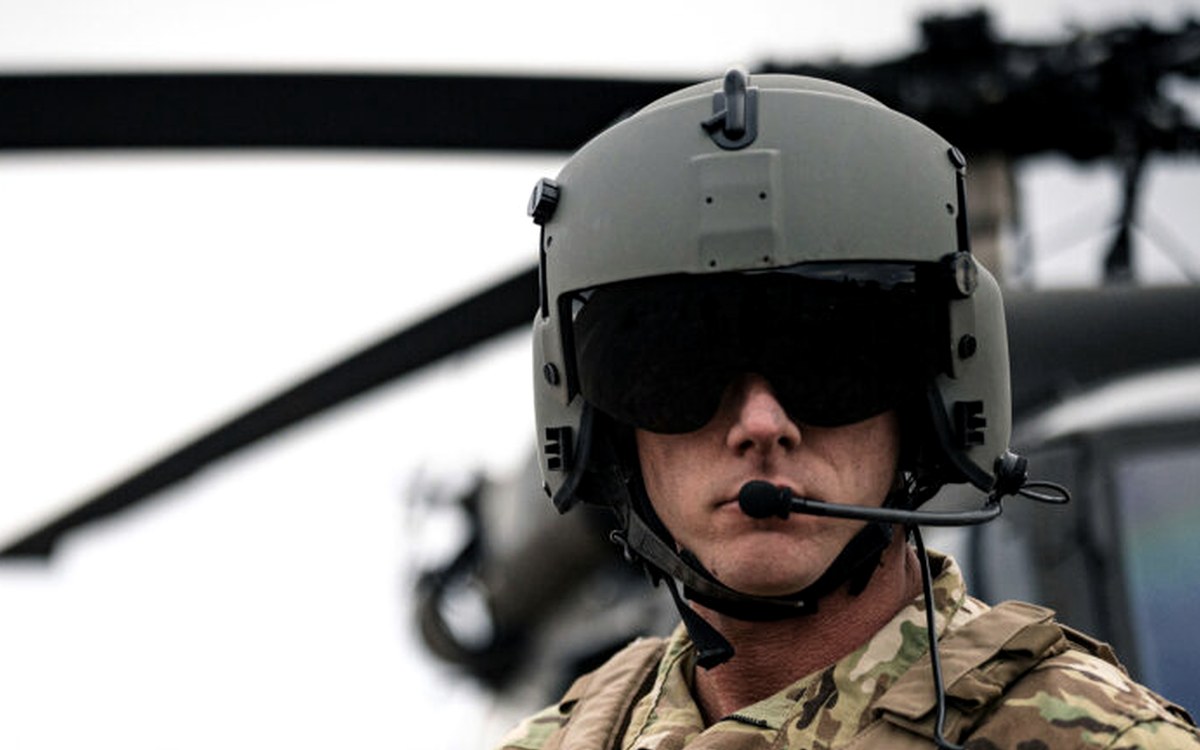 Aircrew helmet with boom microphone and hearing-protection ear cups beside a helicopter